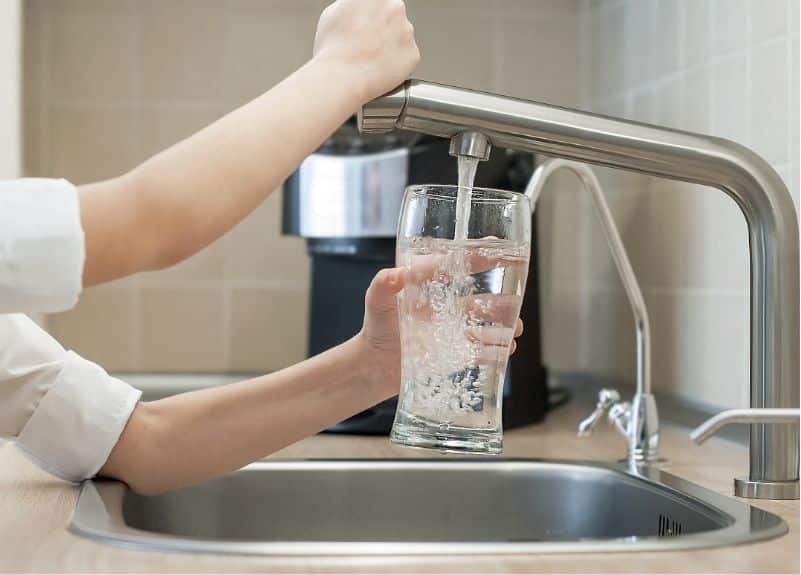 Eine Person füllt ein Glas mit Wasser aus einem modernen Küchenhahn, während eine hochwertige Kaffeemaschine das Ambiente der Küche dezent aufwertet. Die ruhige häusliche Szene bildet mühelos einen Kontrast zum geschäftigen Industriegebiet.