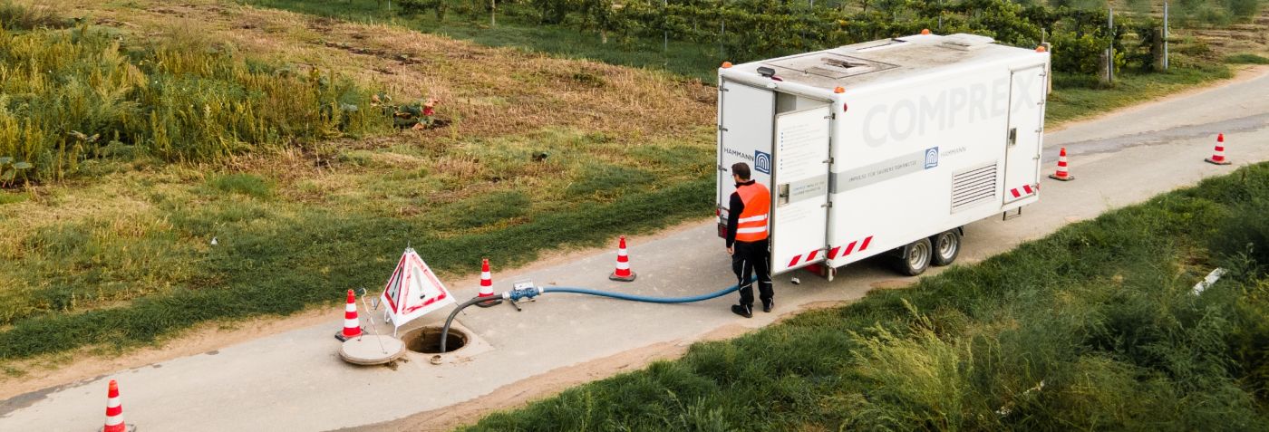 A worker in an orange vest stands next to an open manhole, utilizing Rohrreinigung techniques beside a vehicle and traffic cones on a rural road.