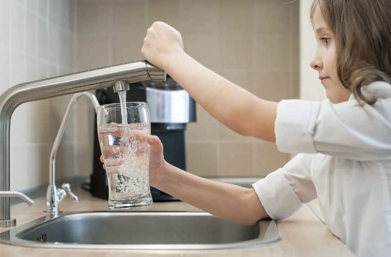 Ein Kind füllt ein Glas mit Wasser aus einem Küchenhahn, Teil des Prozesses „Reinigung von Trinkwasseranlagen“.