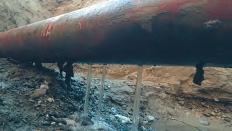 A large, elevated metal pipe is shown leaking water in multiple places into the dirt and rocky ground below, as if neglected after a pig cleaning operation.