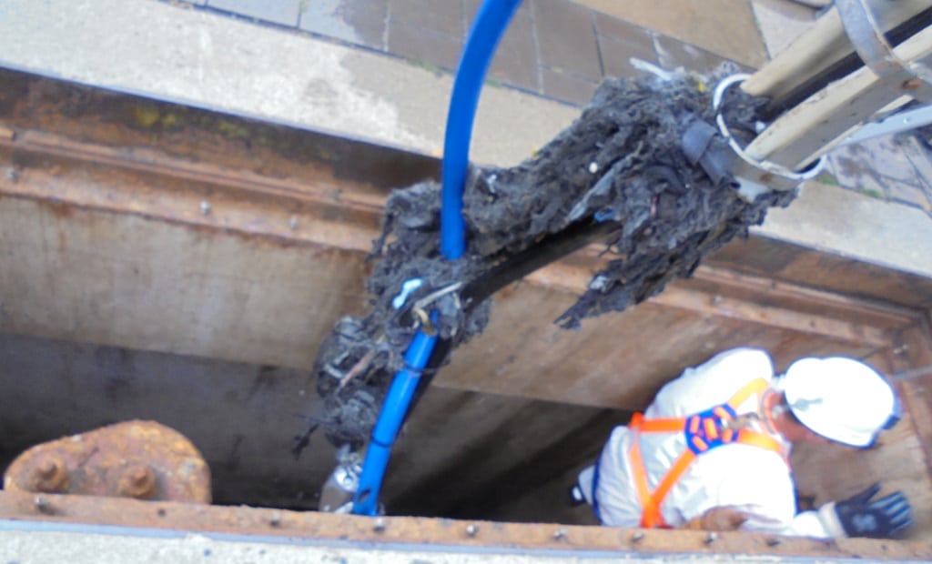 A worker in safety gear is lowered into a sewer through an open manhole, examining a tool entangled with debris and organic matter attached to blue cables.