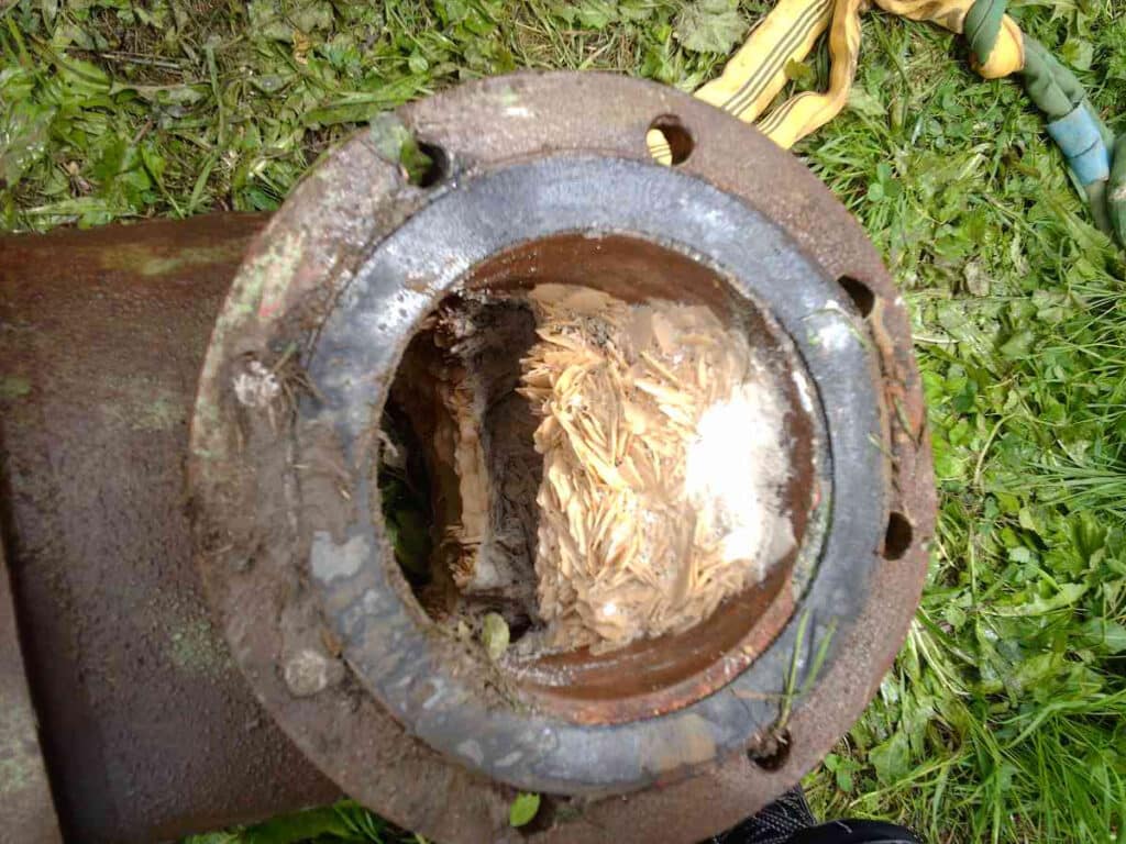 A close-up image of a rusted pipe section with a buildup of fibrous material blocking the interior, indicative of the need for pig cleaning, lying on a grassy surface.