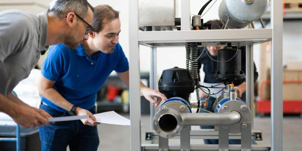 Two men examine machinery with pipes and equipment in an industrial setting, possibly discussing pipeline and plant cleaning with comprex®. One holds a paper, while the other points at a component.