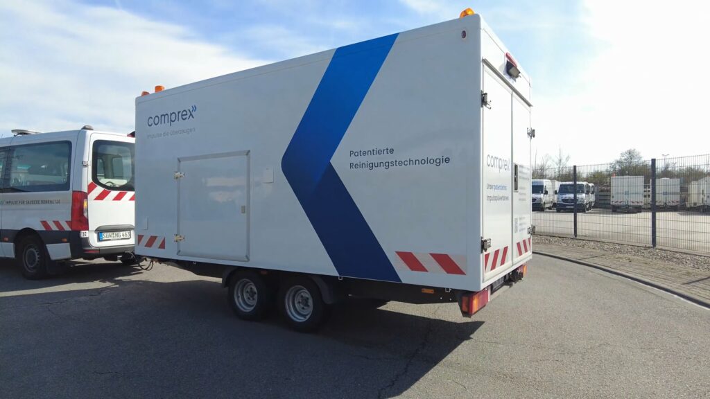 Weißer Anhänger mit Aufschrift „Comprex® patentierte Reinigungstechnologie“, geparkt auf einer Asphaltfläche, blauer Himmel über ihm, demonstriert fortschrittliche Rohrleitungs- und Anlagenreinigung mit Comprex®.