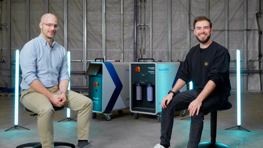 Zwei Männer sitzen in einer Lagerhalle mit Industrieanlagen dazwischen und demonstrieren ihr Fachwissen in der Rohrleitungs- und Anlagenreinigung mit Comprex®. Beide lächeln in die Kamera.